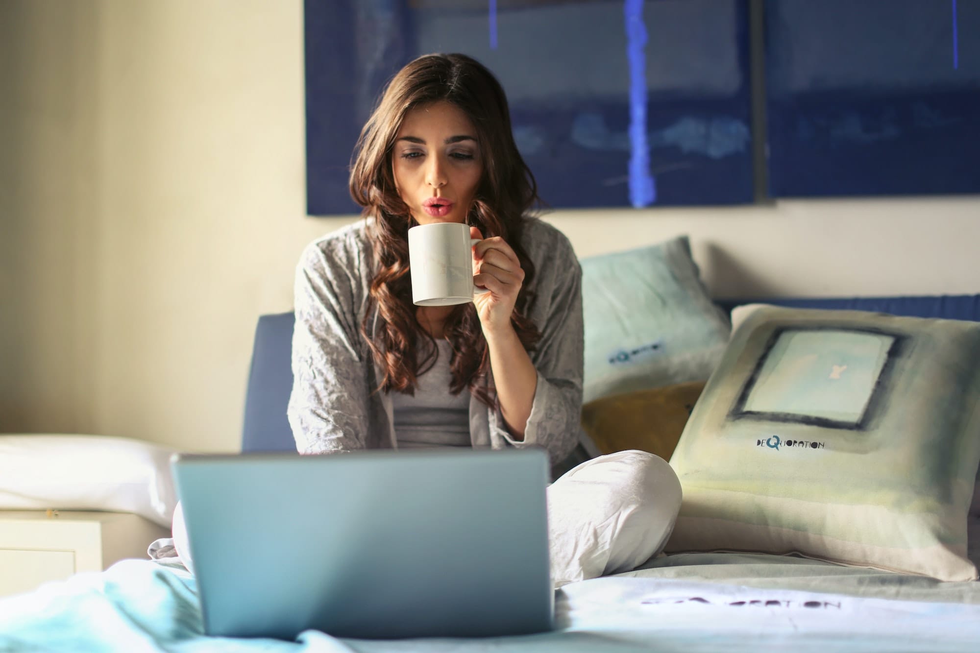 A woman drinking from a mug on her bed in her apartment.